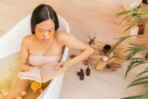 A woman enjoys a relaxing bath with eye patches, reading a book surrounded by skincare items.